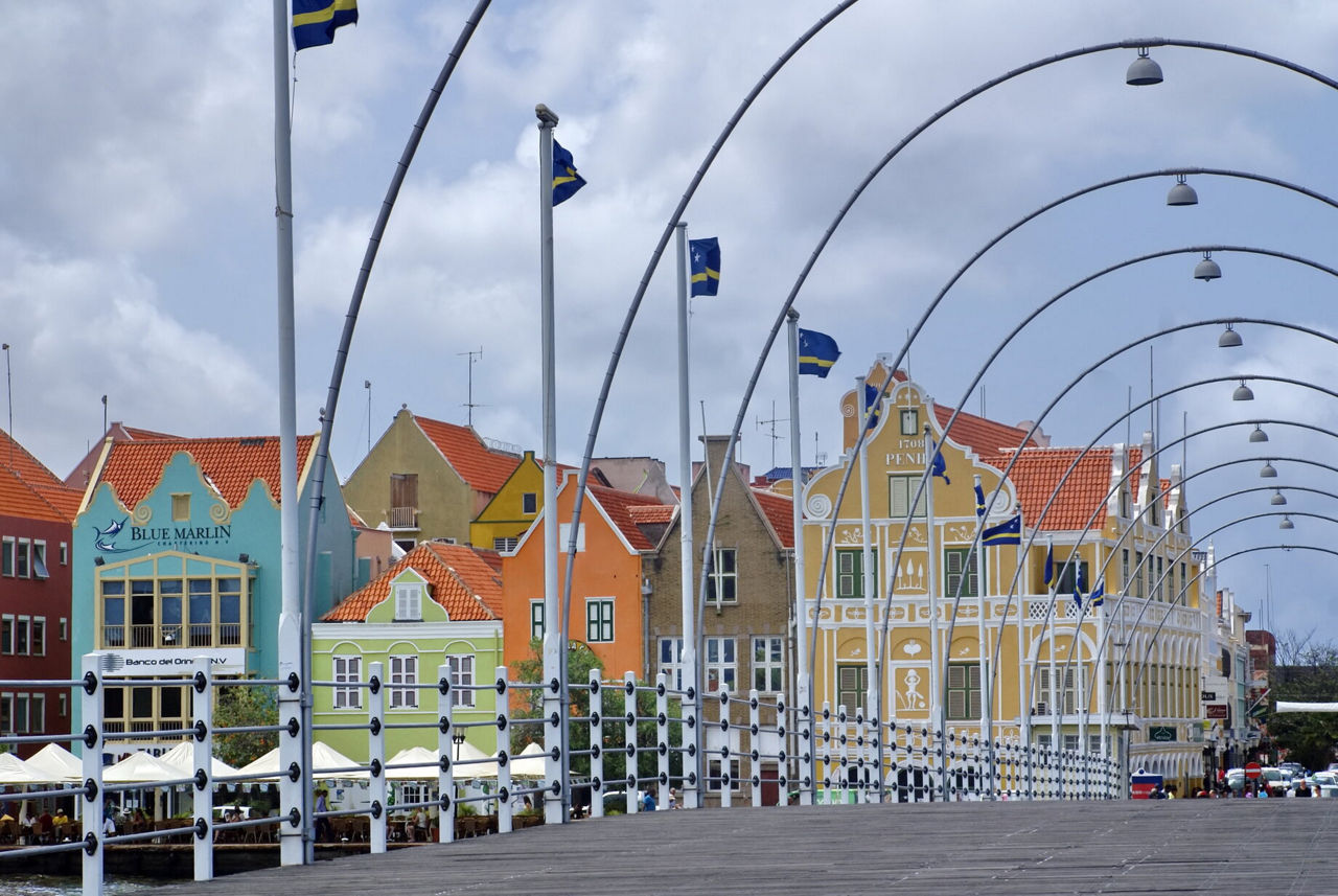 The Queen Emma Bridge in Willemstad, Curaçao, featuring arched metal supports, waving Curaçao flags and a backdrop of vibrant Dutch colonial buildings with terracotta roofs.