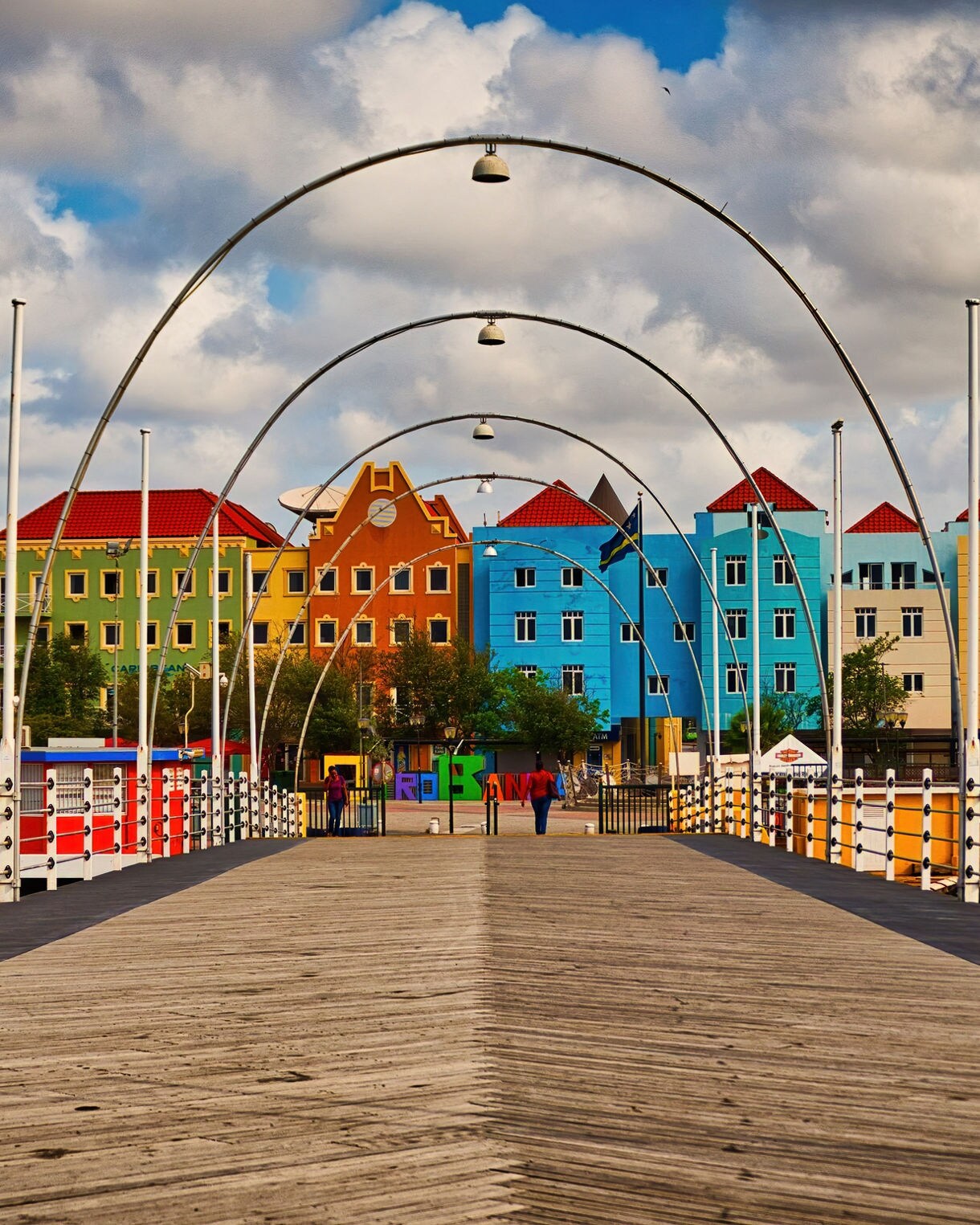 The Queen Emma Bridge in Willemstad, Curaçao, featuring wooden planks and arched metal frames, with colorful Dutch colonial buildings lining the waterfront under a partly cloudy sky.