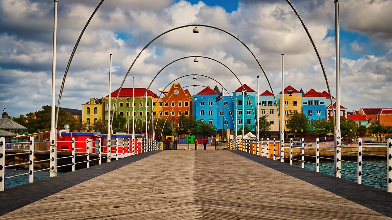 The Queen Emma Bridge in Willemstad, Curaçao, featuring wooden planks and arched metal frames, with colorful Dutch colonial buildings lining the waterfront under a partly cloudy sky.