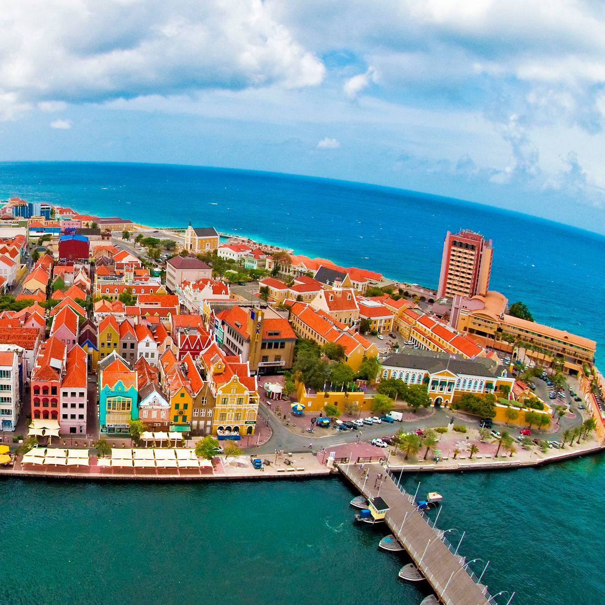 Aerial view of Willemstad's peninsula with red-roofed colorful buildings surrounded by turquoise ocean, boats and a floating pontoon bridge.