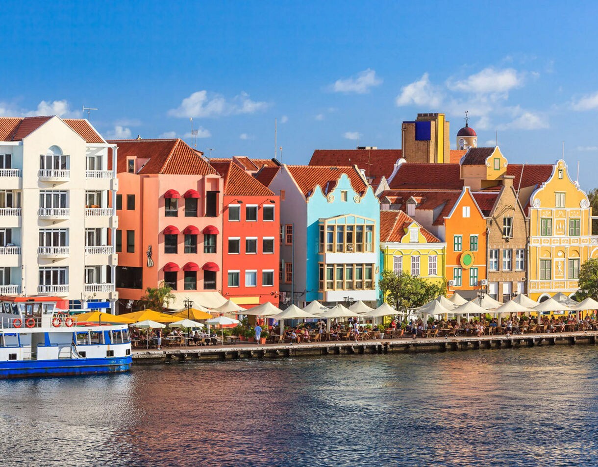 A vibrant row of Dutch colonial buildings with red-tiled roofs along the waterfront in Willemstad, Curaçao, featuring outdoor cafés with white umbrellas and a blue ferry docked nearby.