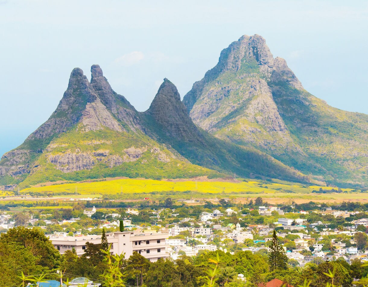 Tall jagged mountains rising behind a town with fields and trees in the foreground.