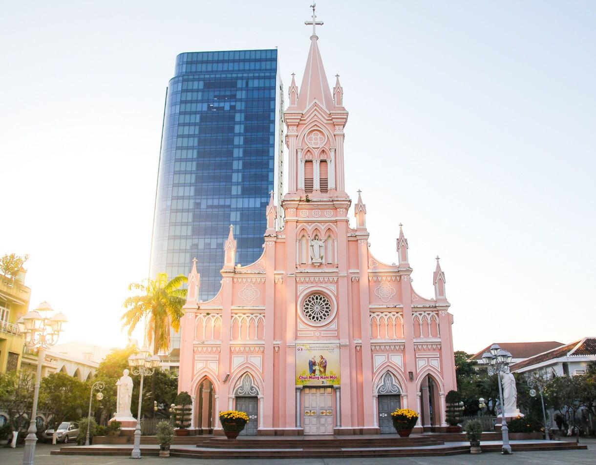 Front view of Da Nang Cathedral, a pastel pink Gothic-style church with arched windows and a tall spire, set against a modern glass skyscraper in the background.