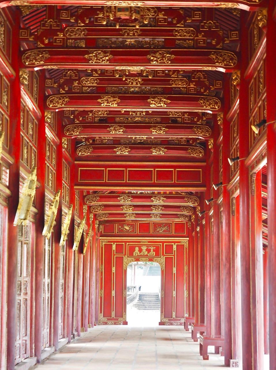 Interior hallway of the Dai Noi Royal Palace in Hue, Vietnam, featuring deep red columns and intricately carved gold details leading to a bright doorway.
