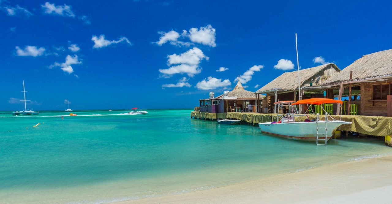 A tropical beach scene with bright turquoise water, white sand, and wooden buildings with thatched roofs built over the water. Several boats are moored nearby, including a white boat with an orange canopy. Sailboats can be seen in the distance under a brilliant blue sky with white clouds.