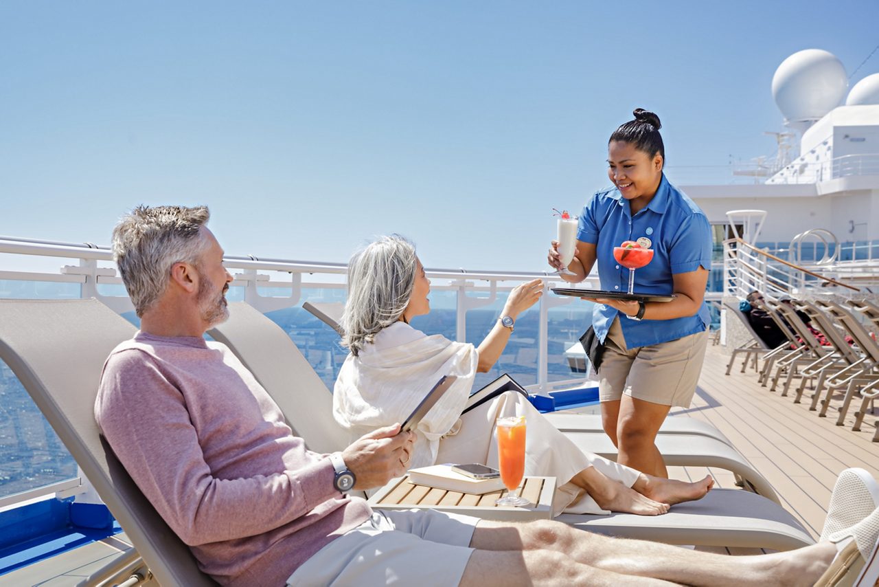 A smiling server in a blue uniform hands a tropical drink to a woman with gray hair sitting on a lounge chair on a deck of a ship. Next to her, a man in a pink sweater reclines on another lounge chair, smiling and holding a menu. A small table with a drink and menus is in the foreground, with a clear blue sky and ocean visible in the background.