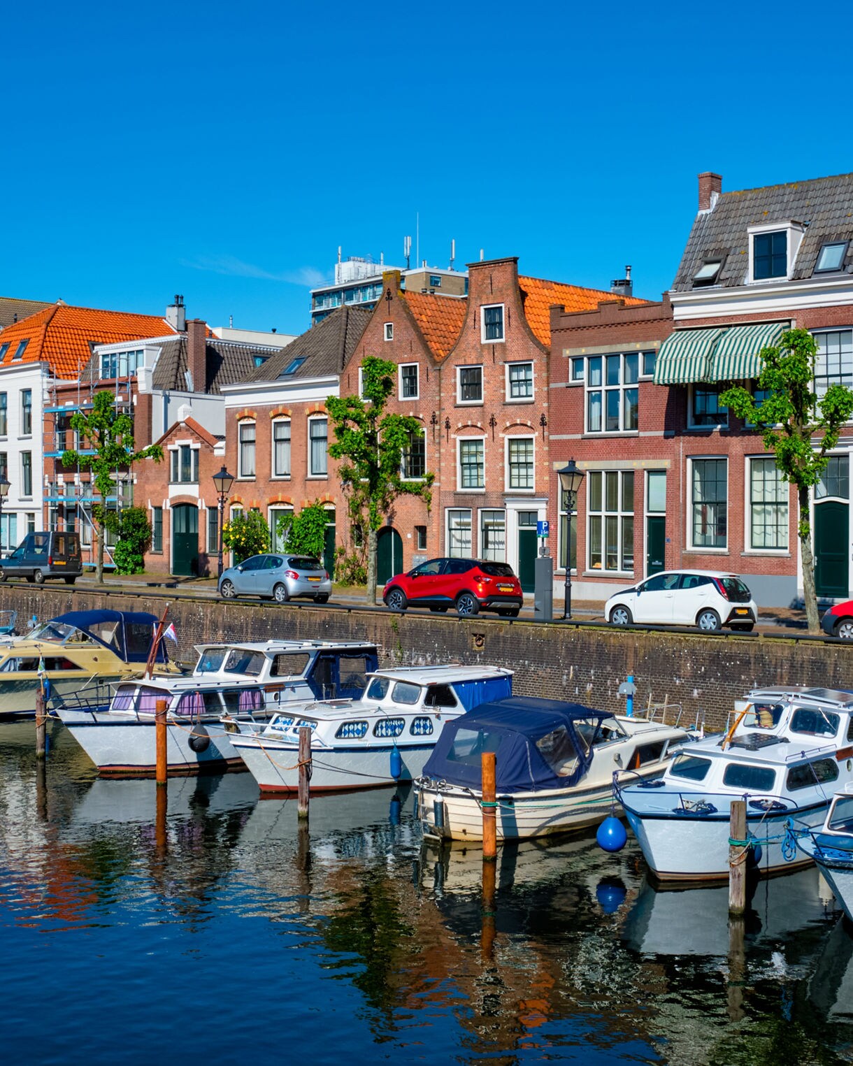 Delfshaven harbor lined with small boats in calm water, fronted by historic brick houses with gabled roofs and bright window details under a clear blue sky.