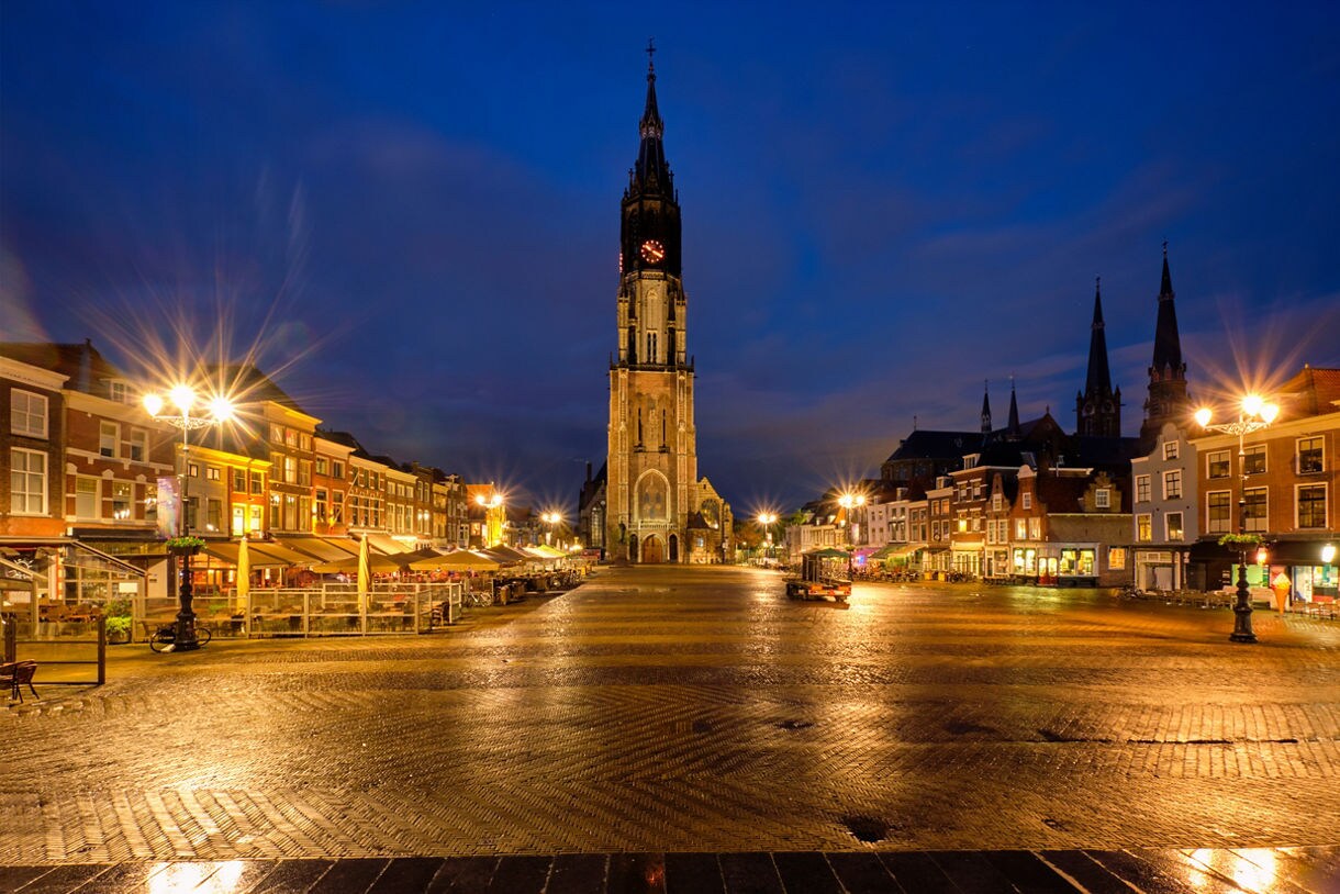 Delft’s Markt square at night with the illuminated Nieuwe Kerk church in the center, flanked by colorful historic buildings and outdoor cafes under warm streetlights.