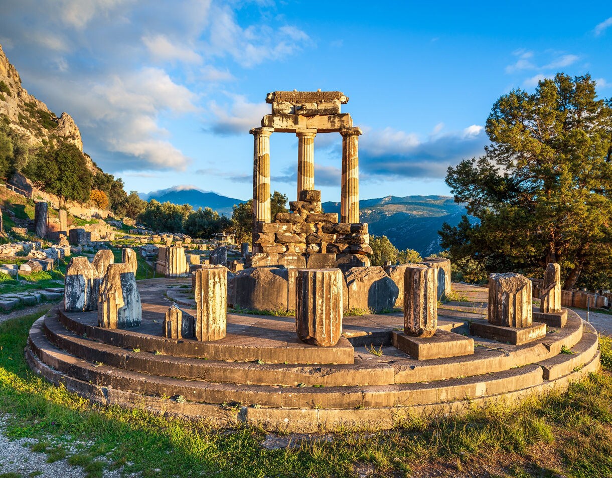 The circular ruins of the Tholos at Delphi, featuring a few standing columns and scattered stone remains, set against rugged mountains and a bright sky.
