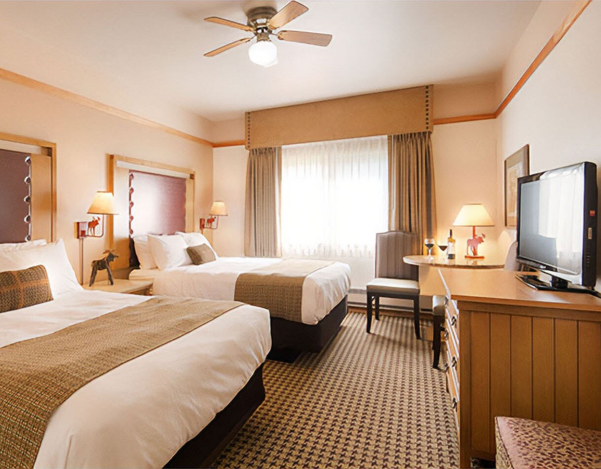 Well-appointed hotel room with two queen beds dressed in white linens with tan accent runners, warm wood trim, ceiling fan, flat-screen TV, armchair, and natural light filtering through sheer curtains.