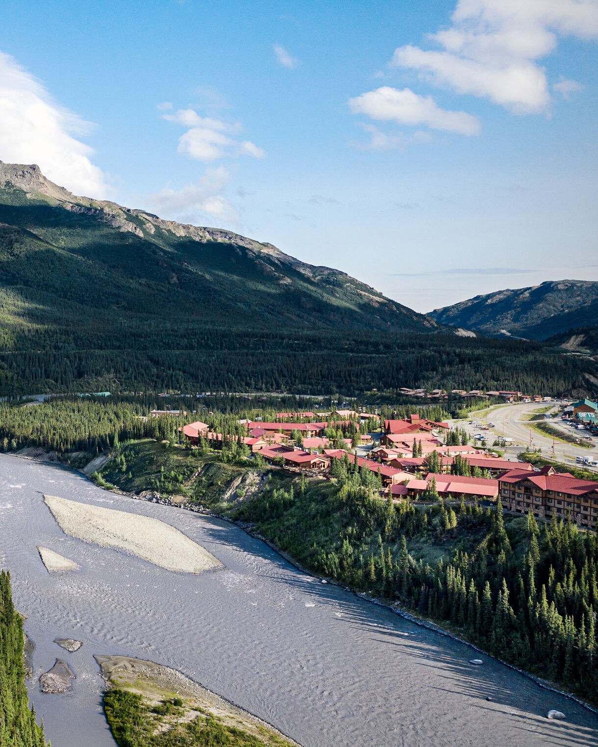 Aerial view of Denali Princess Wilderness Lodge with red-roofed buildings nestled among spruce forest along the Nenana River with forested mountains rising in background under blue sky with white clouds.