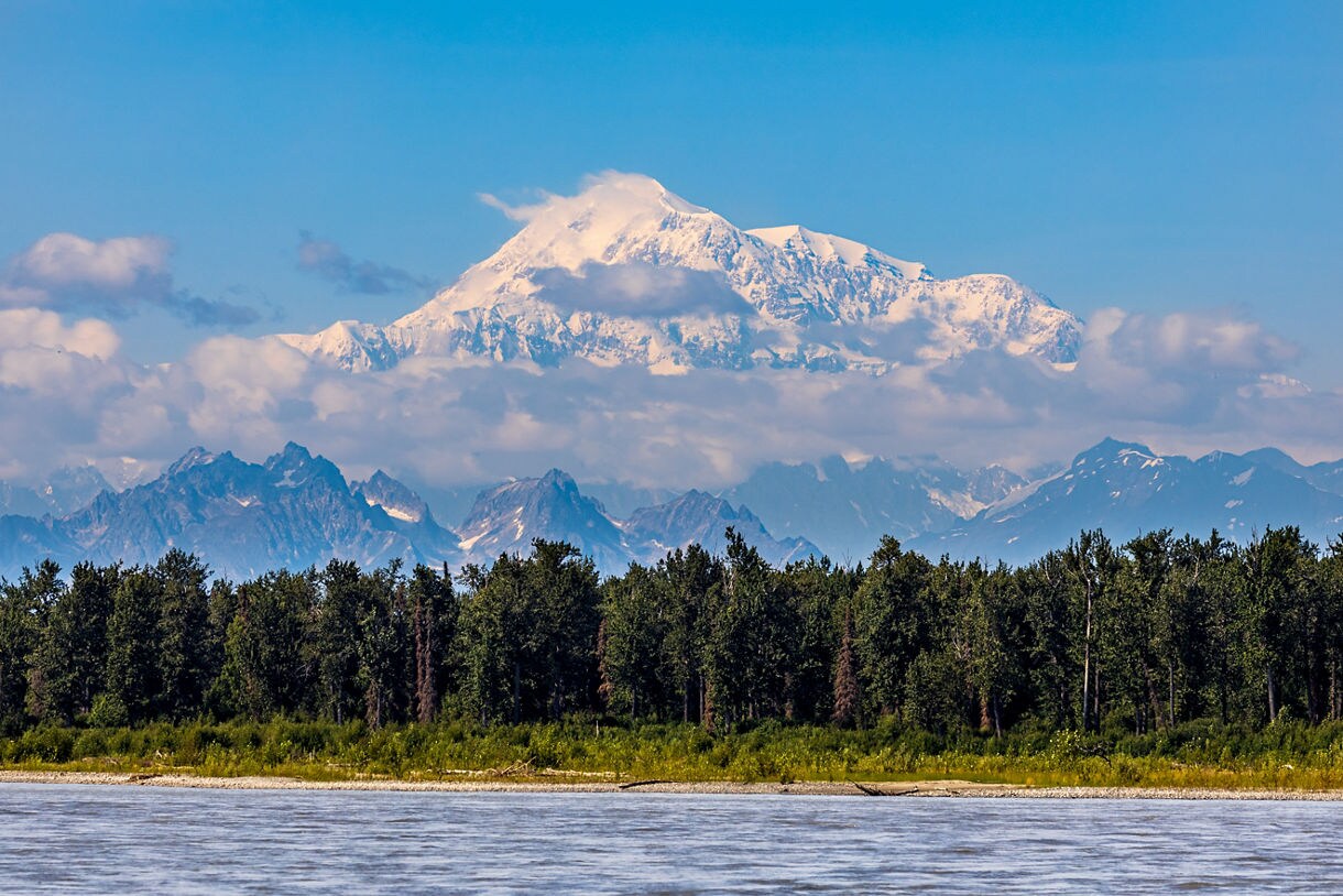 A stunning view of Denali as seen from Talkeetna, Alaska, featuring its snow-capped peak, forest trees, and a calm body of water in the foreground.