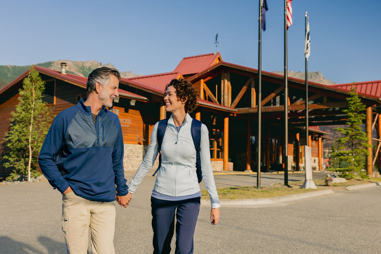  Couple holding hands while walking toward the main lodge building featuring log construction, red metal roof, and flagpoles at Denali Wilderness Lodge
