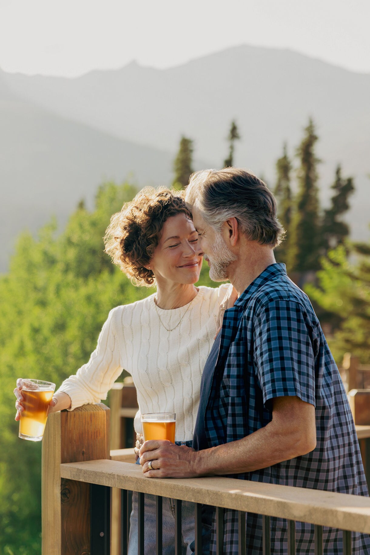 Couple enjoying craft beers together on wooden deck railing with misty mountain landscape and pine trees in soft-focus background at Denali Wilderness Lodge