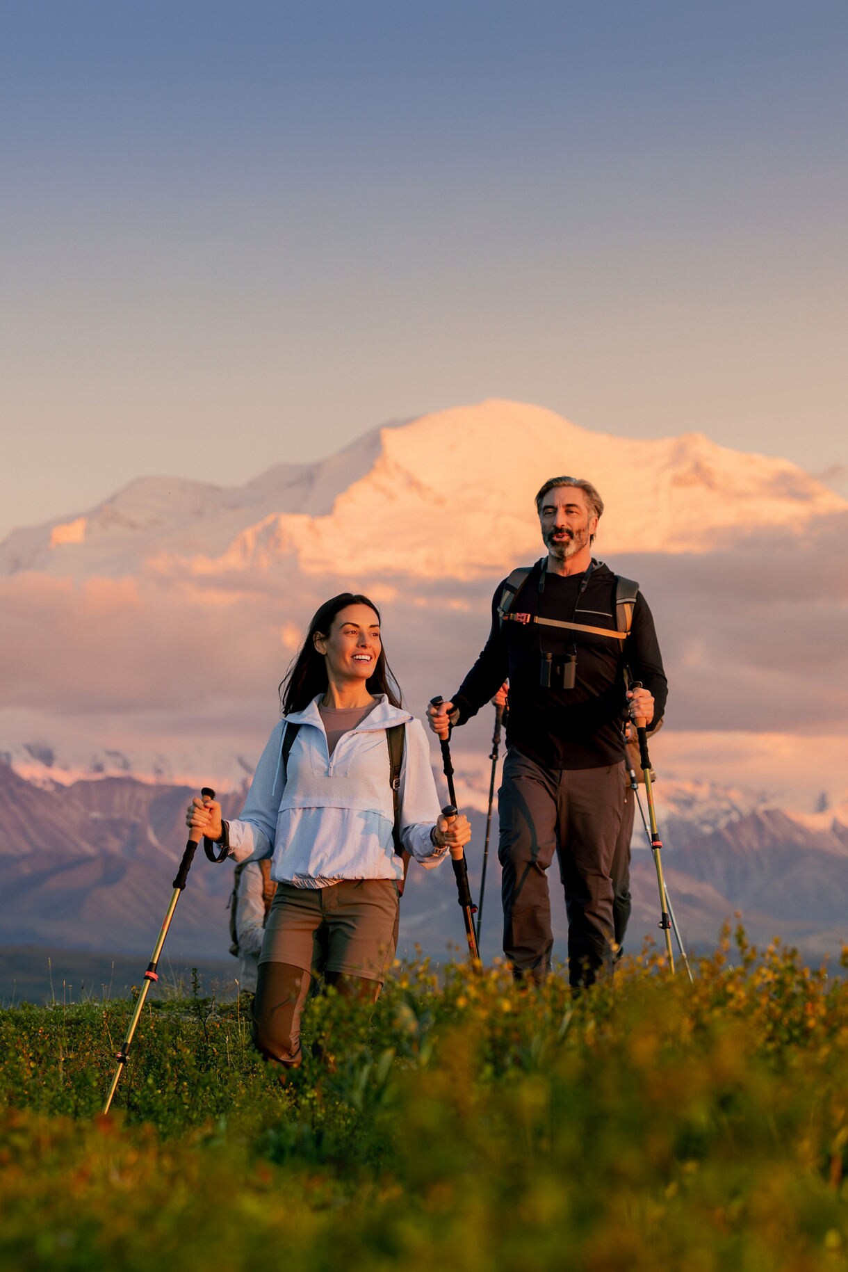 Two hikers with trekking poles standing on alpine meadow at sunset with dramatic mountain vista and golden clouds behind them near Denali Wilderness Lodge