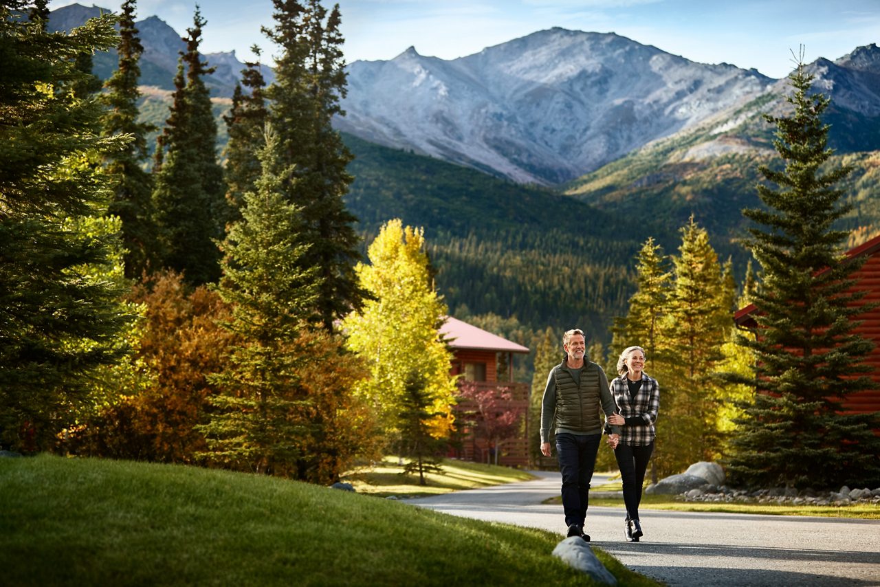 Couple walking on paved path through Denali Princess Wilderness Lodge grounds with colorful autumn spruce trees, lodge cabin visible in background, and snow-capped Alaska Range mountains under clear sky.