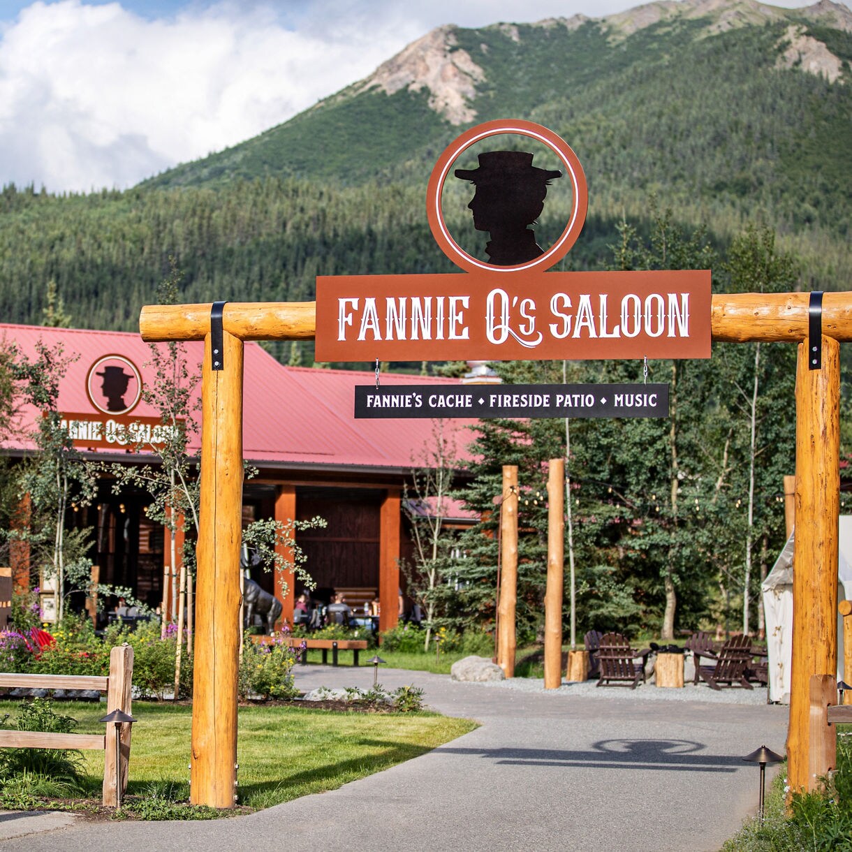 Entrance to Fannie Q's Saloon with large wooden sign displaying cowboy hat logo and text reading Fannie's Cache, Fireside Patio, Music, with pink building and mountain backdrop.