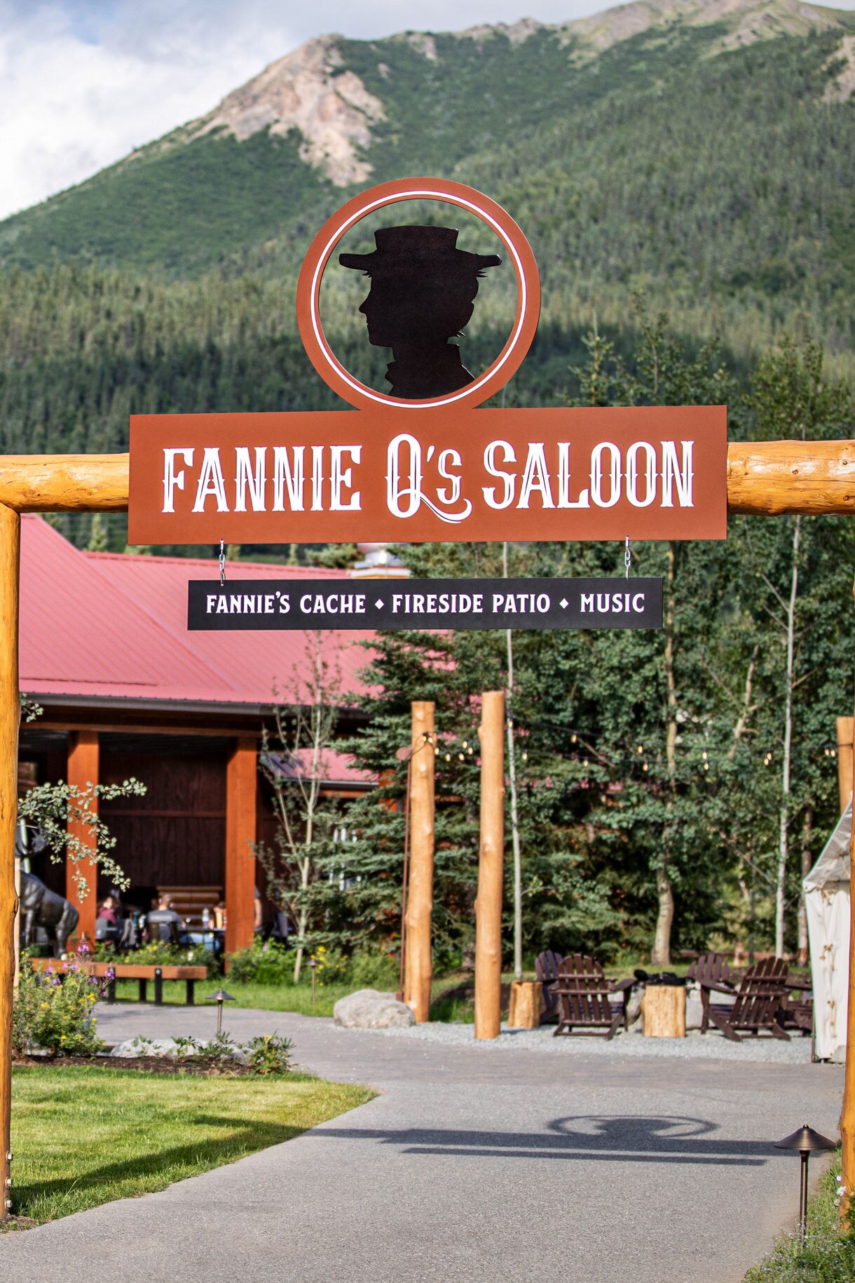  Exterior of Fannie Q's Saloon with rustic wooden signage, fireside patio, and mountain backdrop surrounded by evergreen trees at Denali Wilderness Lodge