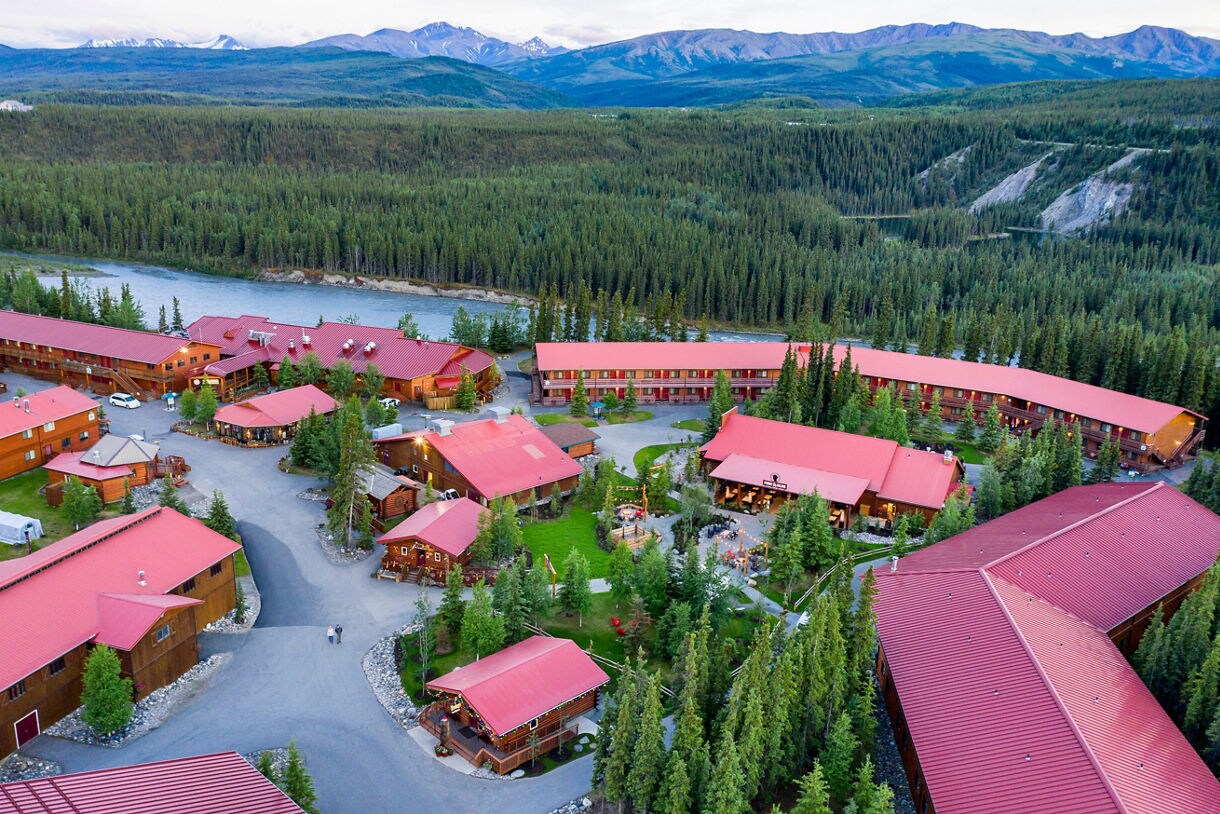  Aerial view of Denali Wilderness Lodge showing multiple buildings with distinctive pink roofs nestled among dense evergreen forest with river and mountain range in background