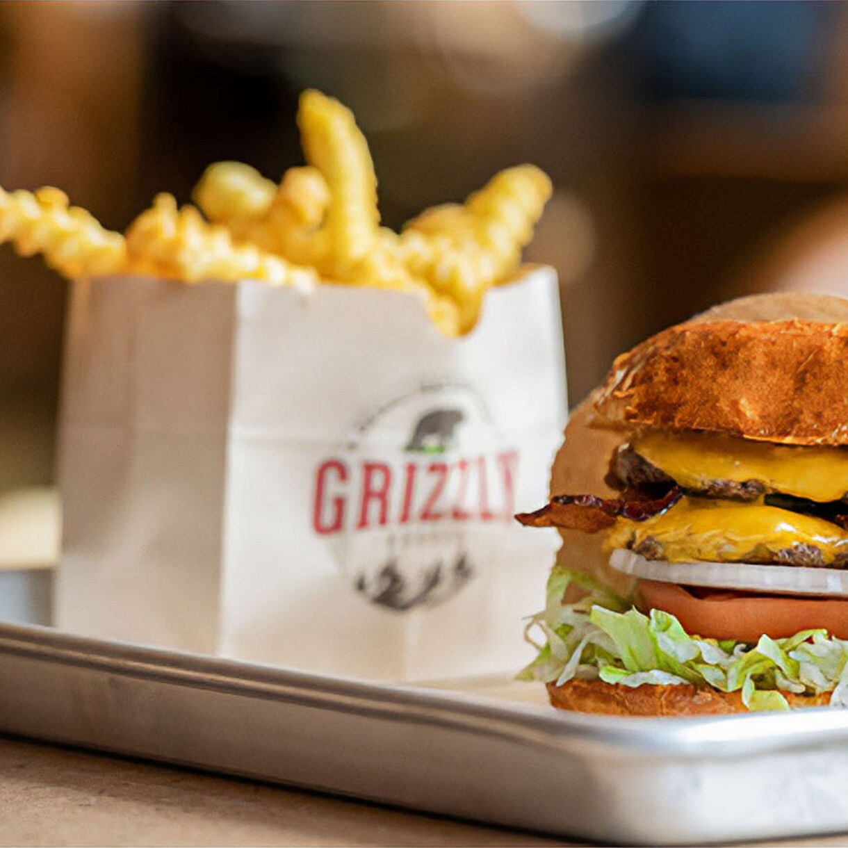 A cheeseburger with bacon, lettuce, tomato, and onion is served on a metal tray alongside crinkle-cut fries in a branded Grizzly bag. A beverage in a clear cup with the Grizzly logo is also visible in the background. The setting appears to be a casual dining restaurant with a warm, inviting atmosphere. The focus is on the fresh ingredients and appetizing presentation. The word 'GRIZZLY' is clearly visible on both the fry bag and drink cup.