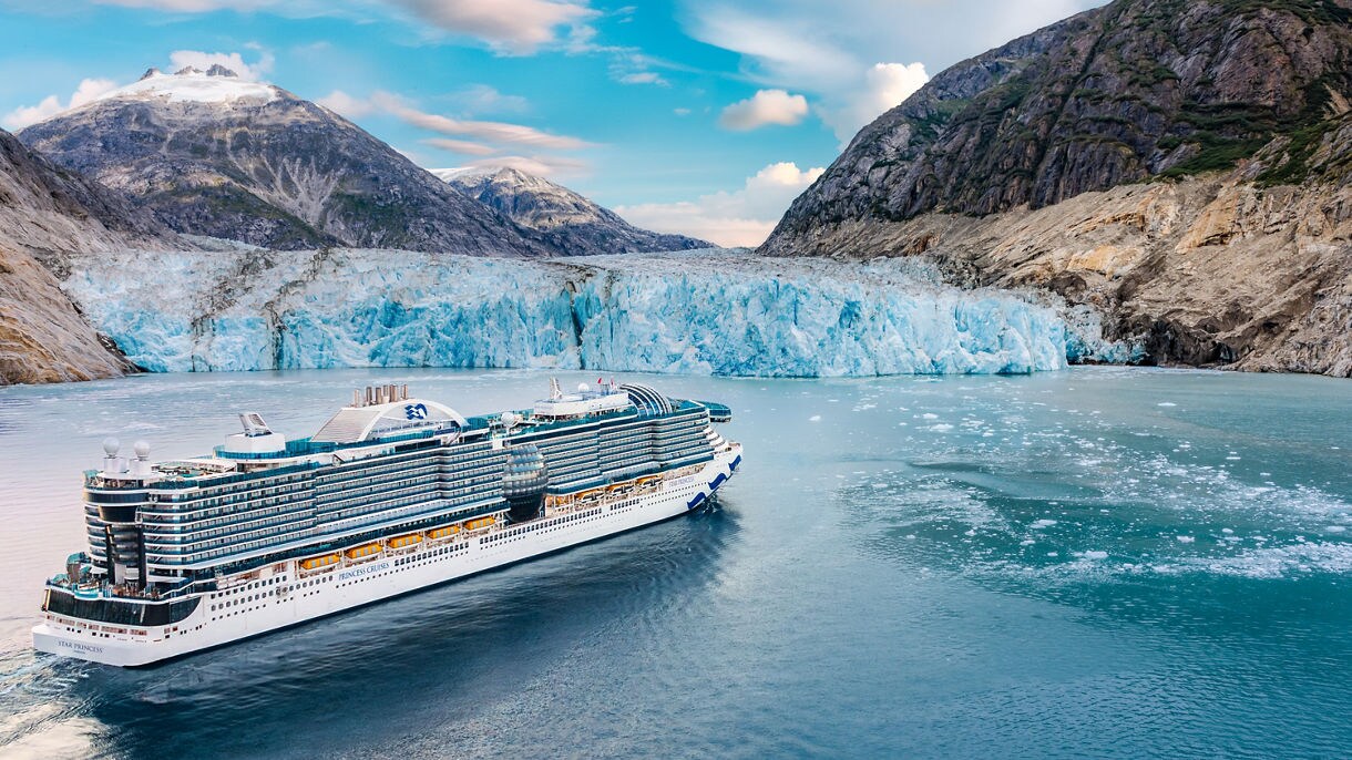 A Princess Cruises ship with distinctive blue and white wave design sails past snow-capped mountains and glaciers in Alaska's waters.