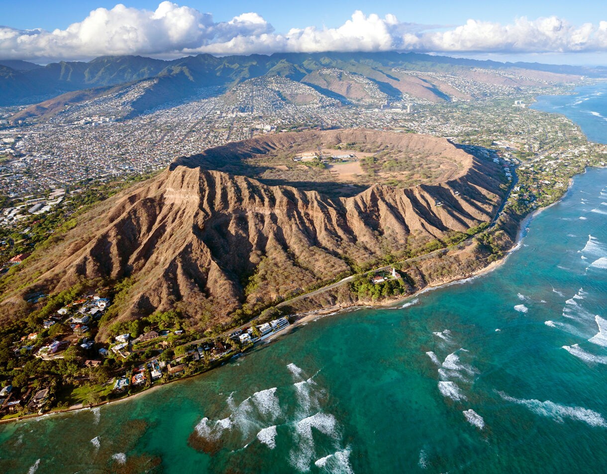 Aerial view of Diamond Head crater in Honolulu, Hawaii, with rugged ridges and turquoise ocean waves along the coastline.