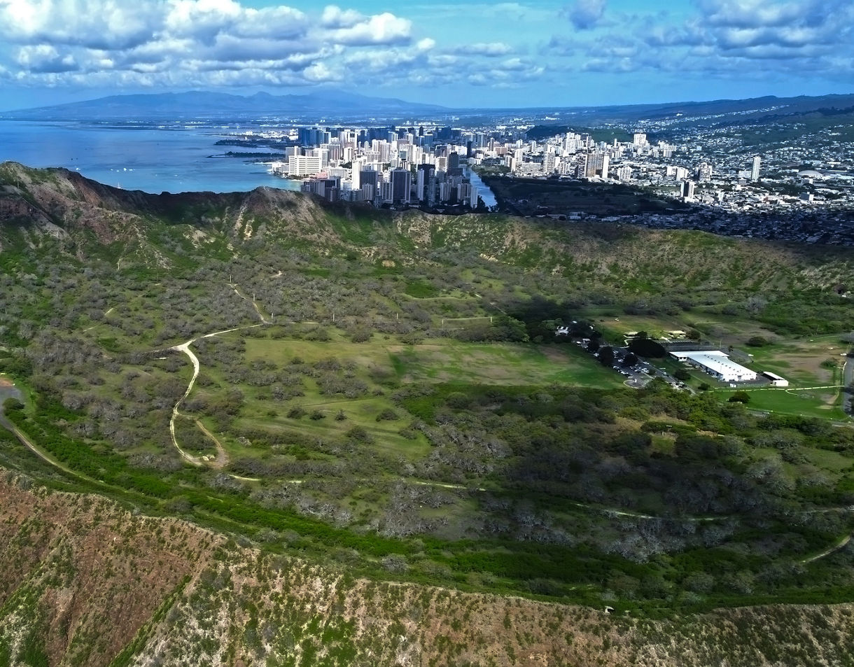 Aerial view of Diamond Head’s crater in Oahu with hiking trails leading to the rim, overlooking Honolulu’s skyline and the blue Pacific beyond.