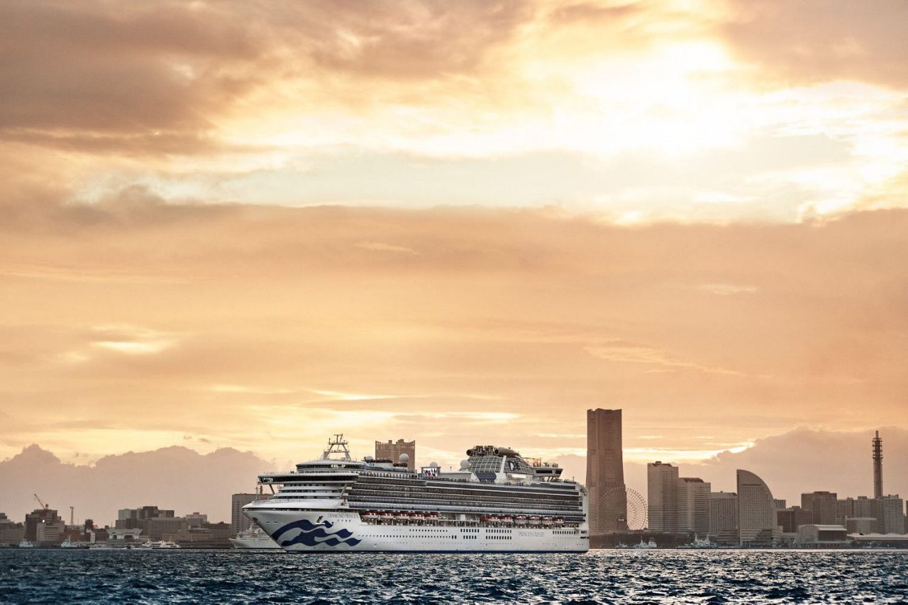 A view of the Diamond Princess cruise ship cruising on calm waters with a city skyline and a warm, golden sunset in the background.