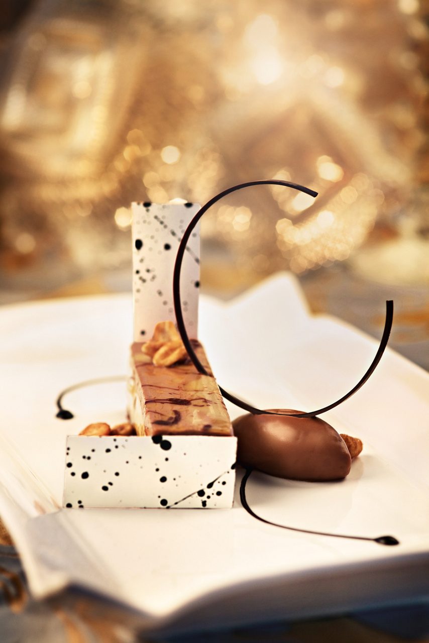 A decadent chocolate peanut butter bar served with a chocolate garnish and a smooth, rich accompaniment, artfully arranged on a white plate with intricate chocolate accents.