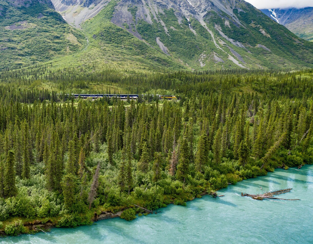 Aerial view of a Princess Cruises train traveling through a vast evergreen forest alongside a turquoise glacial river with mountains in the distance
