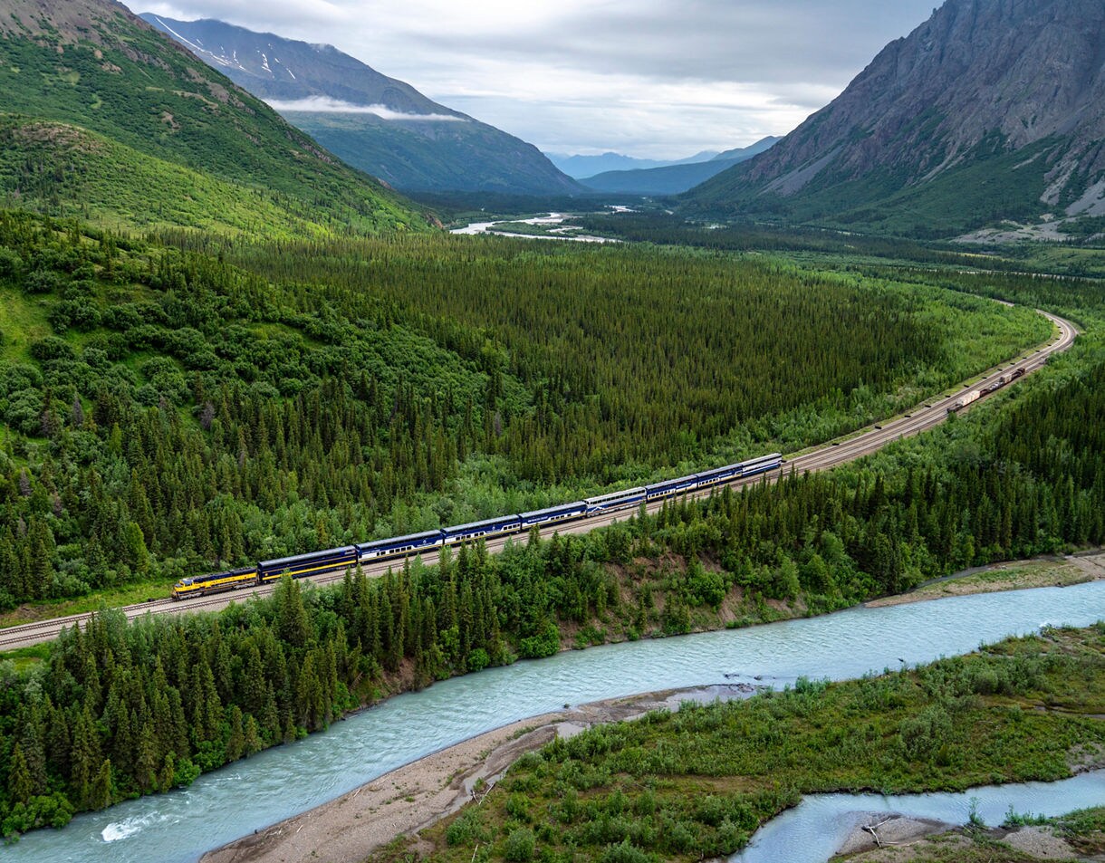 Aerial landscape view of a Princess Cruises train traveling through a mountain valley with evergreen forests, a winding river, and dramatic peaks under cloudy skies