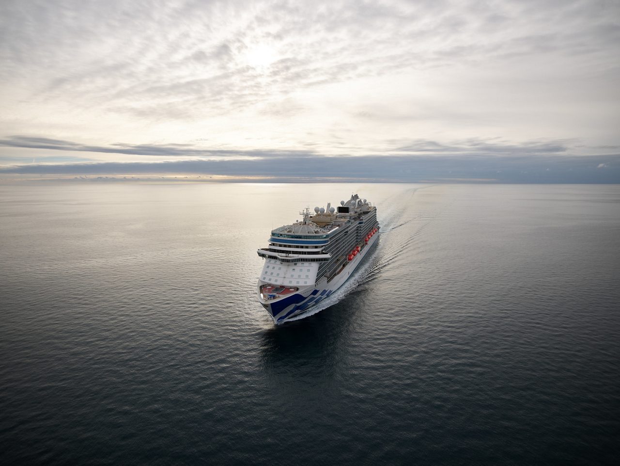 Large white cruise ship with blue accents sails through calm waters, leaving a trail of ripples, with a city skyline of tall buildings and skyscrapers in the background under a vibrant blue and yellow sky. 