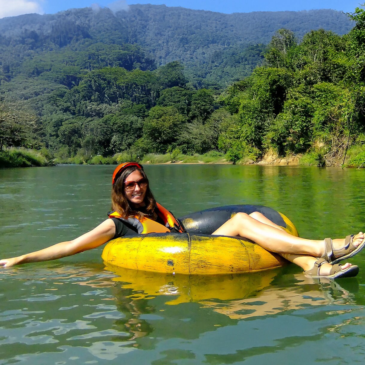 A person tubing on Dominica's Layou River, with clear water, tropical trees lining the banks and mountains in the background. The person is wearing a life jacket and enjoying the serene environment.