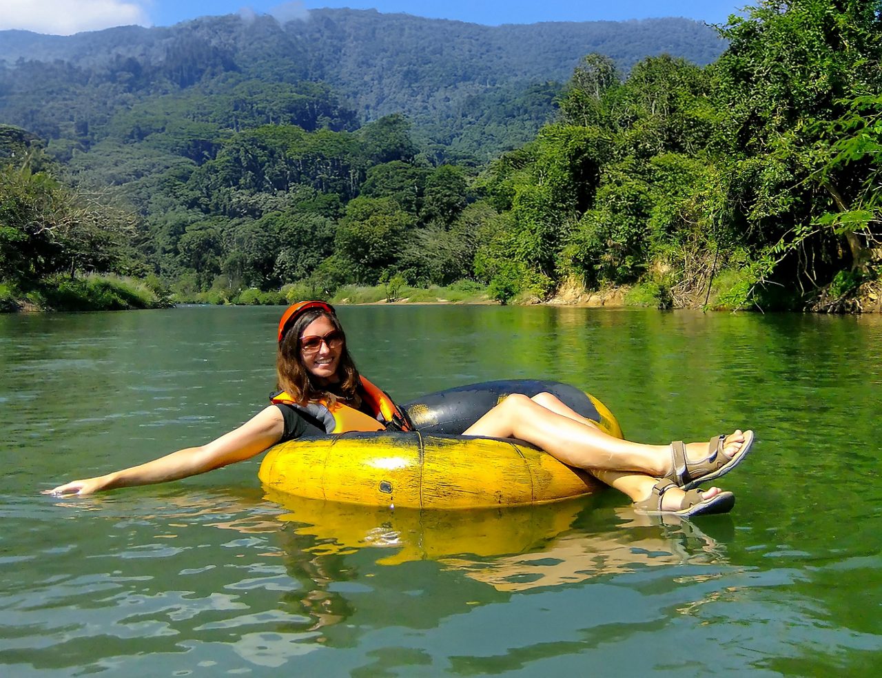 A person tubing on Dominica's Layou River, with clear water, tropical trees lining the banks and mountains in the background. The person is wearing a life jacket and enjoying the serene environment.