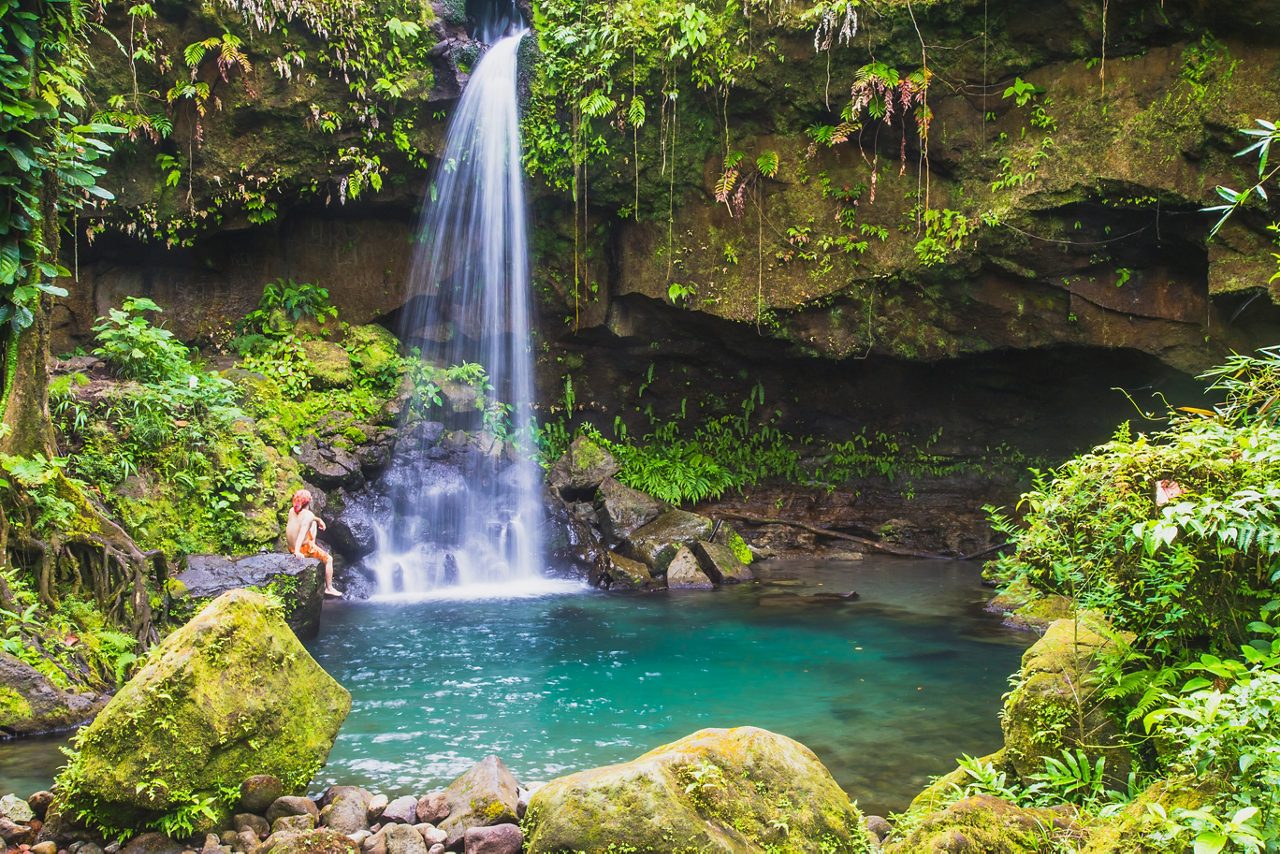 A picturesque waterfall flows into a crystal-clear pool surrounded by vibrant green foliage in Dominica's Emerald Pool, with a visitor sitting on a rock nearby.