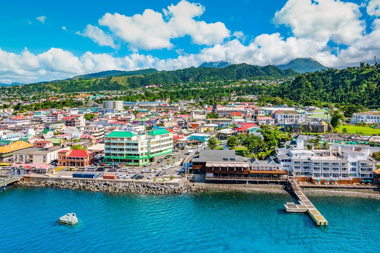 Aerial view of Roseau, Dominica's capital city, showing colorful buildings, a waterfront pier, and mountains in the background.