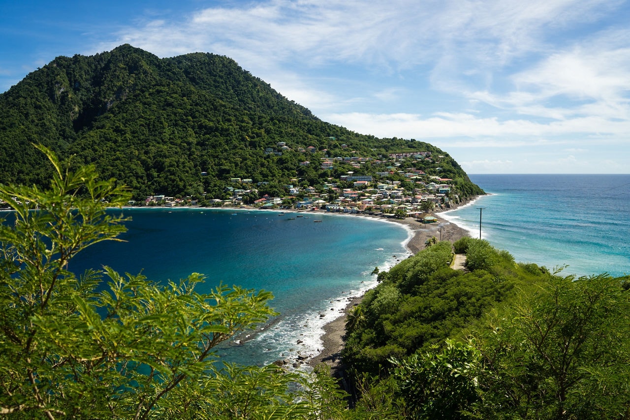Aerial view of Scotts Head Peninsula in Dominica, featuring a lush green hillside, a small coastal village with colorful homes and a narrow strip of land separating the Caribbean Sea from the Atlantic Ocean.