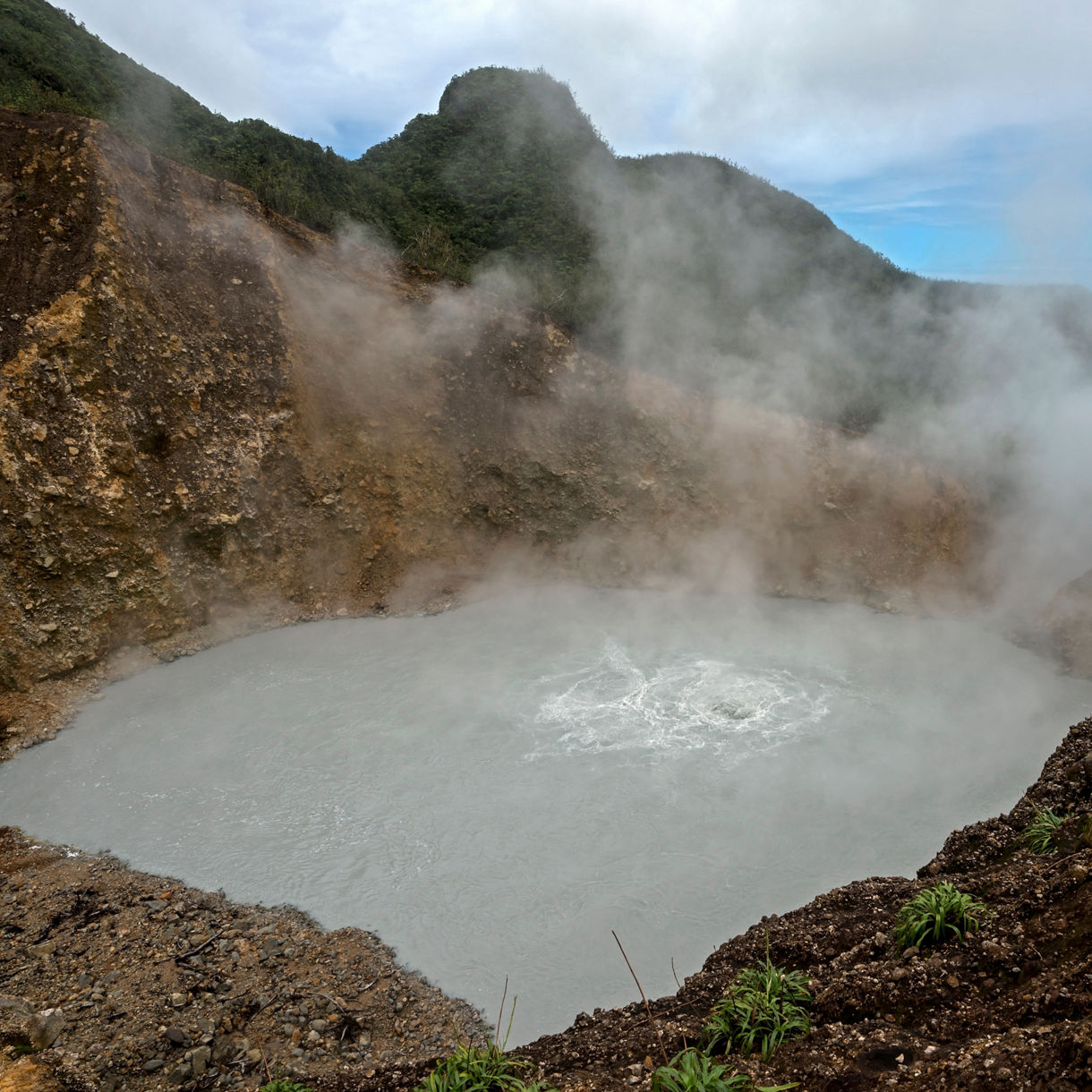 A person walks through a geothermal area, surrounded by rising steam from vents in the rocky ground, with lush green mountains in the background.