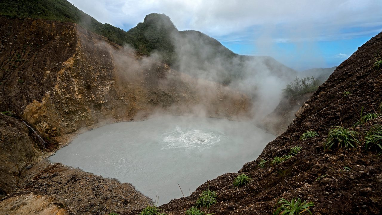 A person walks through a geothermal area, surrounded by rising steam from vents in the rocky ground, with lush green mountains in the background.