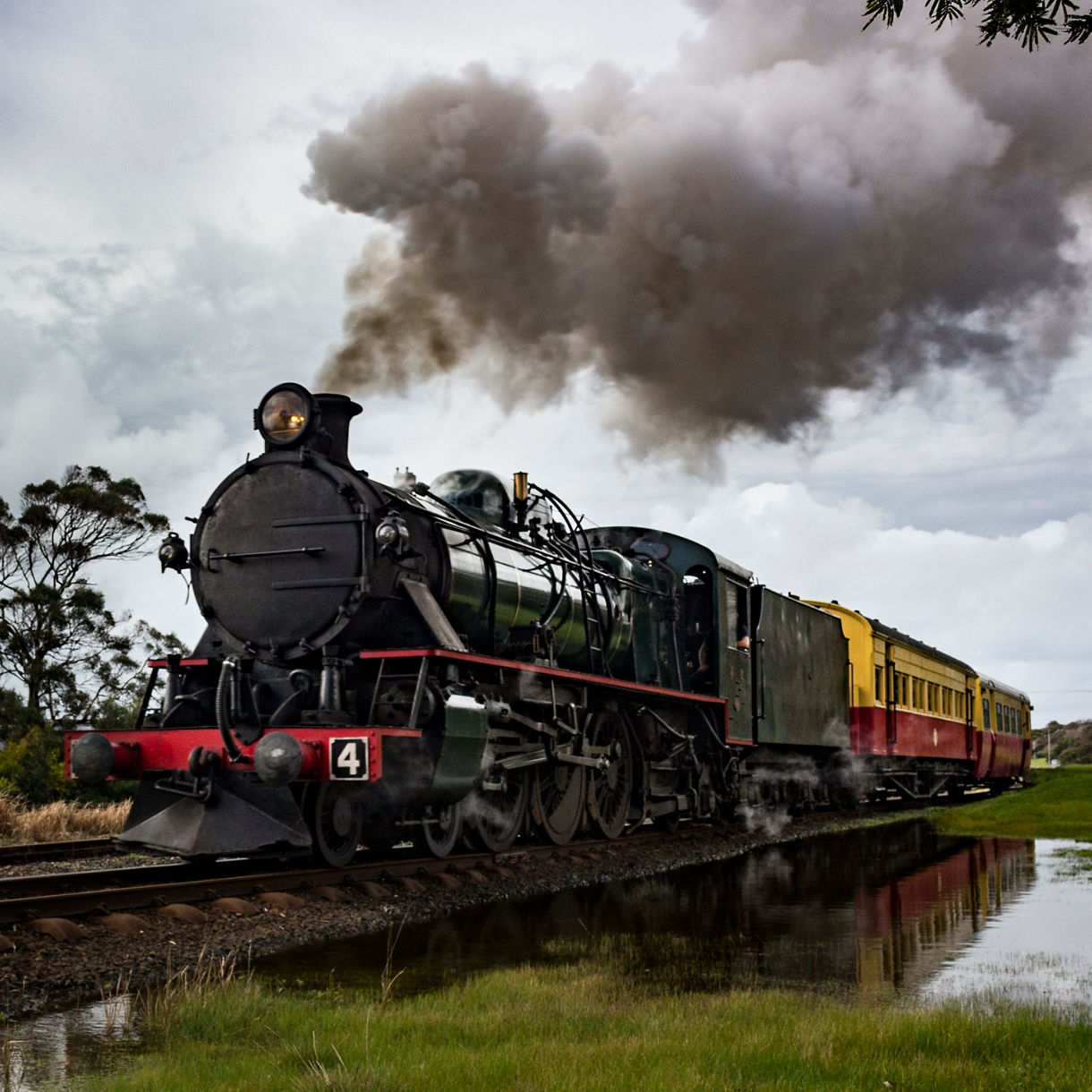 A vintage black steam locomotive pulling yellow and red passenger cars, releasing thick smoke as it travels along a rural railway.