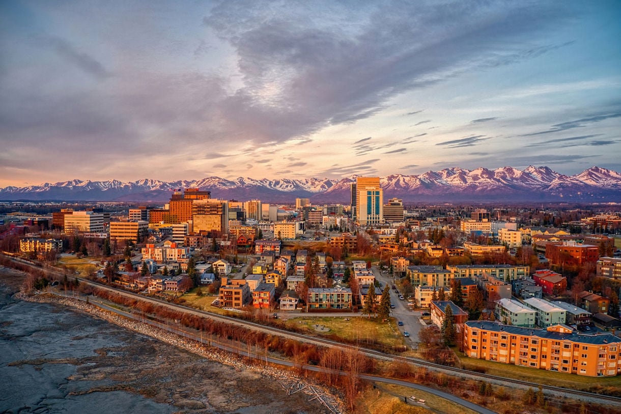 Aerial view of downtown Anchorage, Alaska, during a spring sunset, featuring city buildings, residential areas, and the snow-capped Chugach Mountains illuminated by golden light.