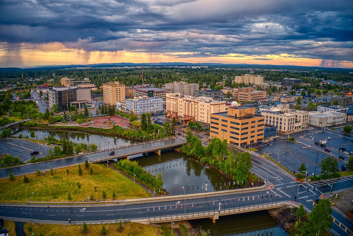 Aerial view of downtown Fairbanks, Alaska, during a summer sunset, showcasing city buildings, bridges, and waterways under dramatic storm clouds with streaks of golden light.