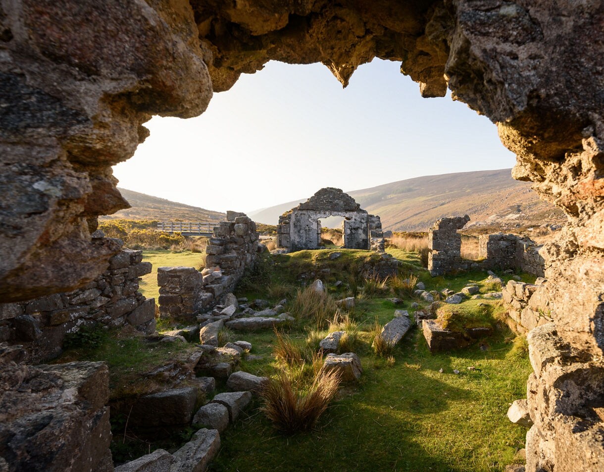 Stone ruins of Glendalough monastery framed by rocky walls, with grassy ground and hills in the background.
