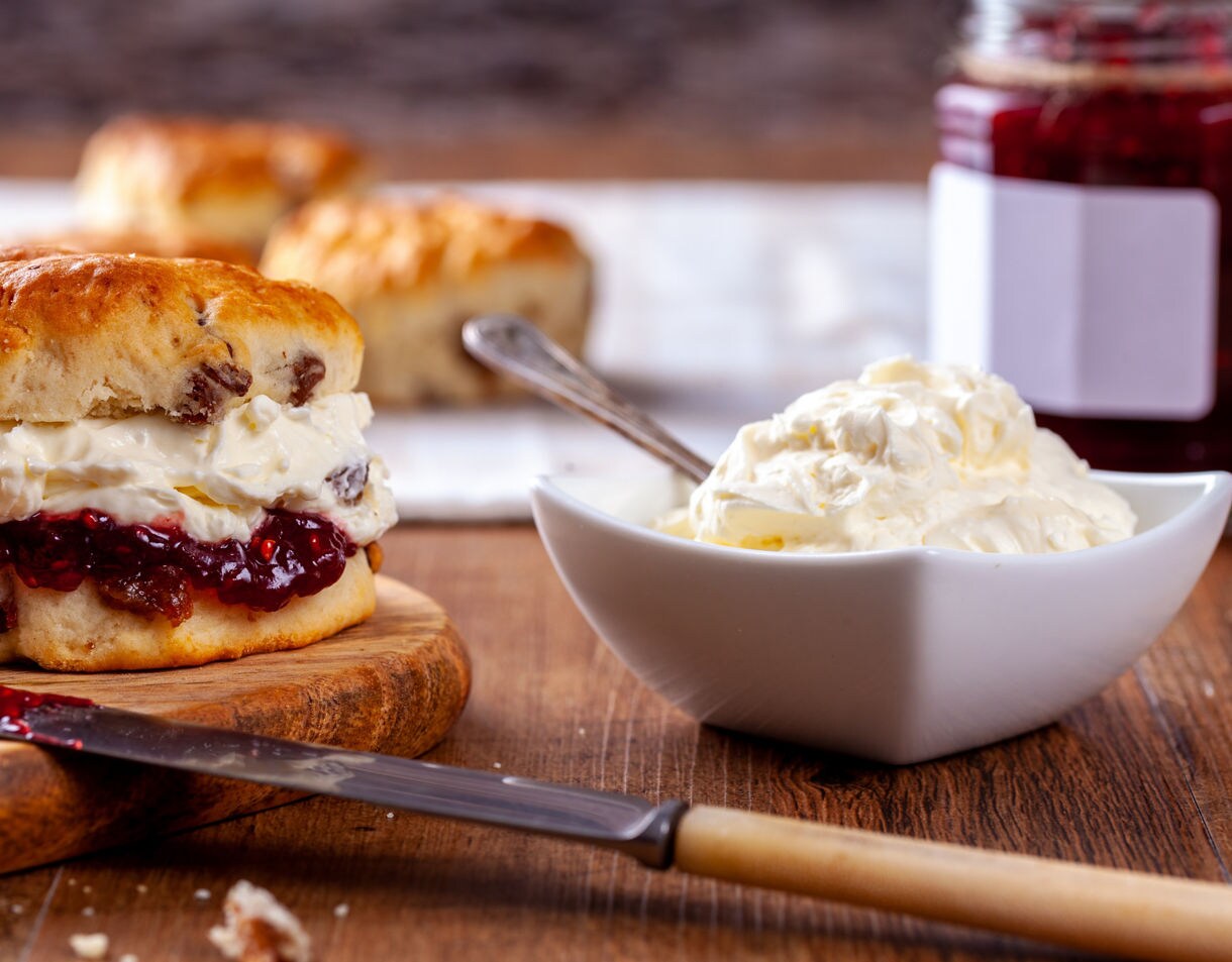 Close-up of a scone filled with clotted cream and raspberry jam, with a bowl of cream and jar of jam in the background.