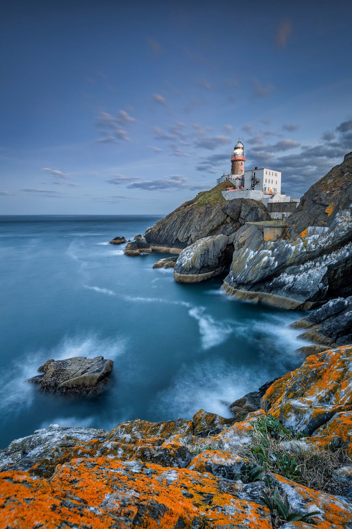 Lighthouse perched on rocky cliffs above the Atlantic Ocean near Dublin, with orange lichen on the foreground rocks.