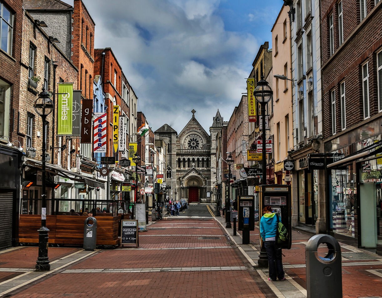 A bustling pedestrian street in Dublin with colorful shop signs and a historic stone church at the end of the road.