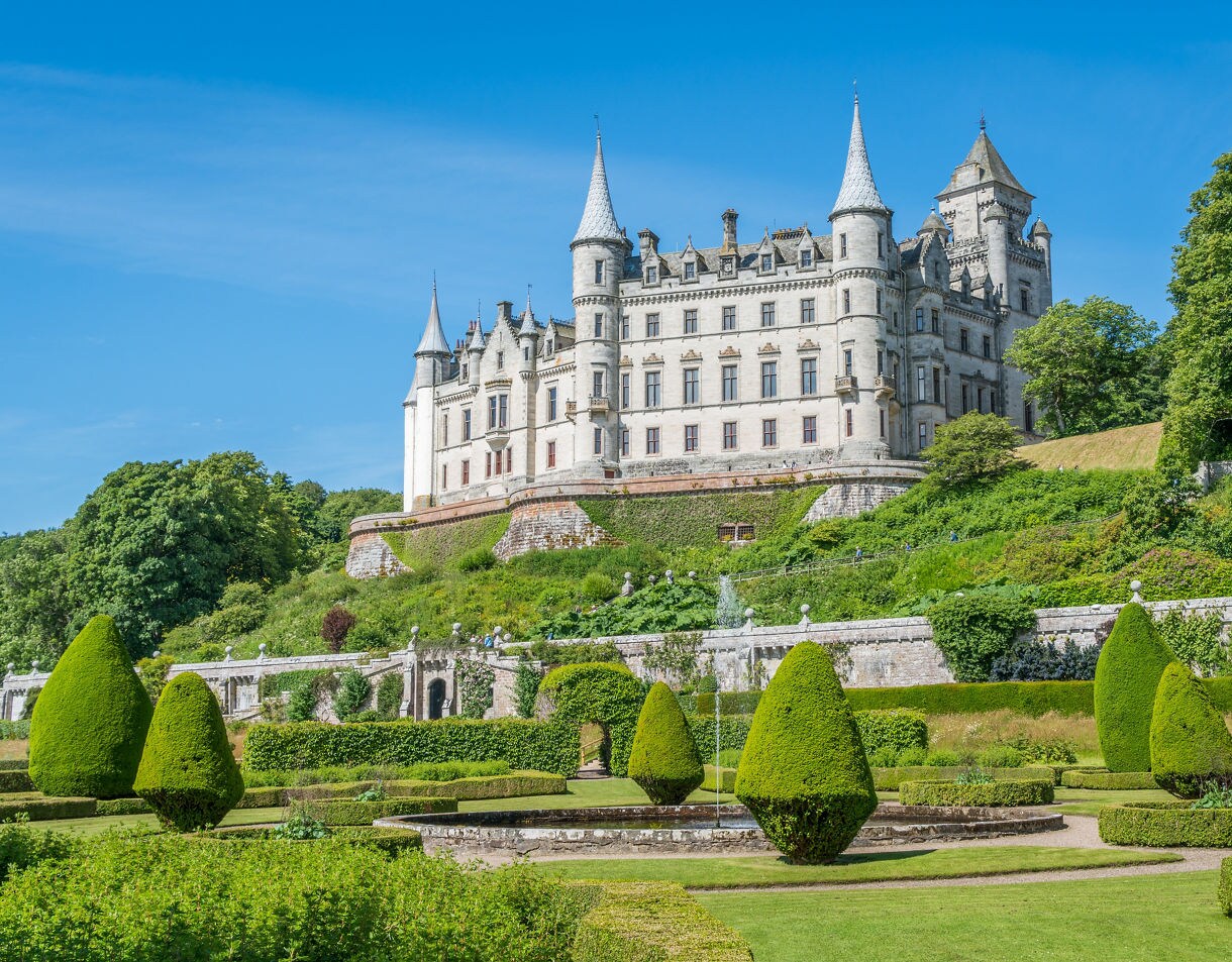 Dunrobin Castle in Scotland, a white château-style estate with pointed spires, perched above formal gardens and trimmed hedges.