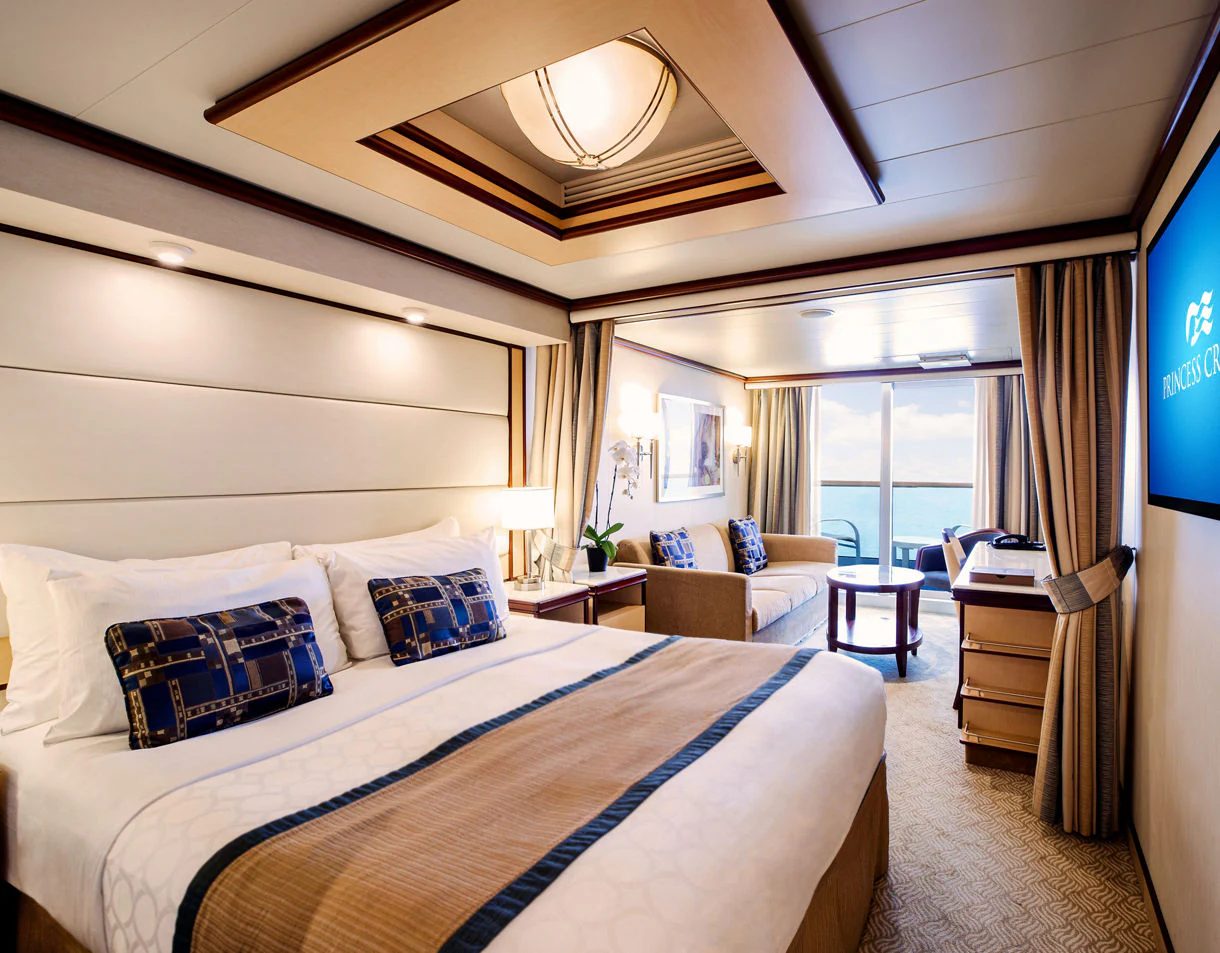  A neatly made double bed with blue and gold accents in a cruise ship stateroom featuring warm wood tones and a window view.