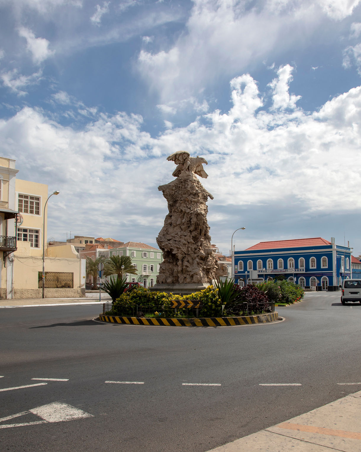 Stone monument topped with an eagle sculpture at the center of a traffic roundabout, surrounded by landscaped plants, pastel and blue colonial buildings and a bright sky filled with soft clouds.