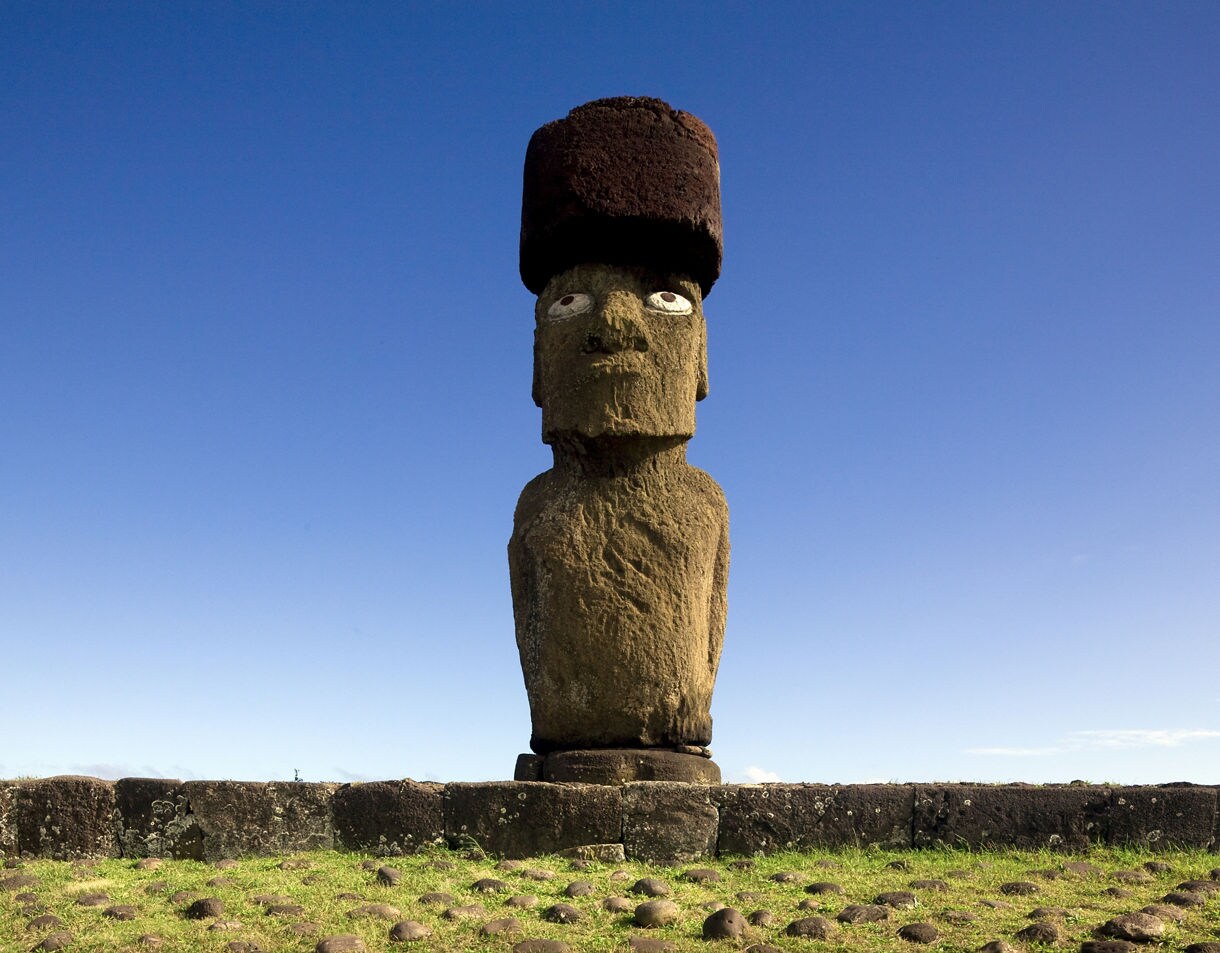 Single moai statue at Ahu Tahai on Easter Island with a red stone topknot and white eyes set against a clear blue sky.